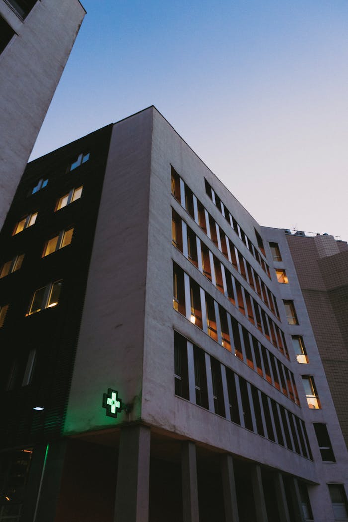 Services-03 Low angle view of a modern hospital building with a green cross at dusk.